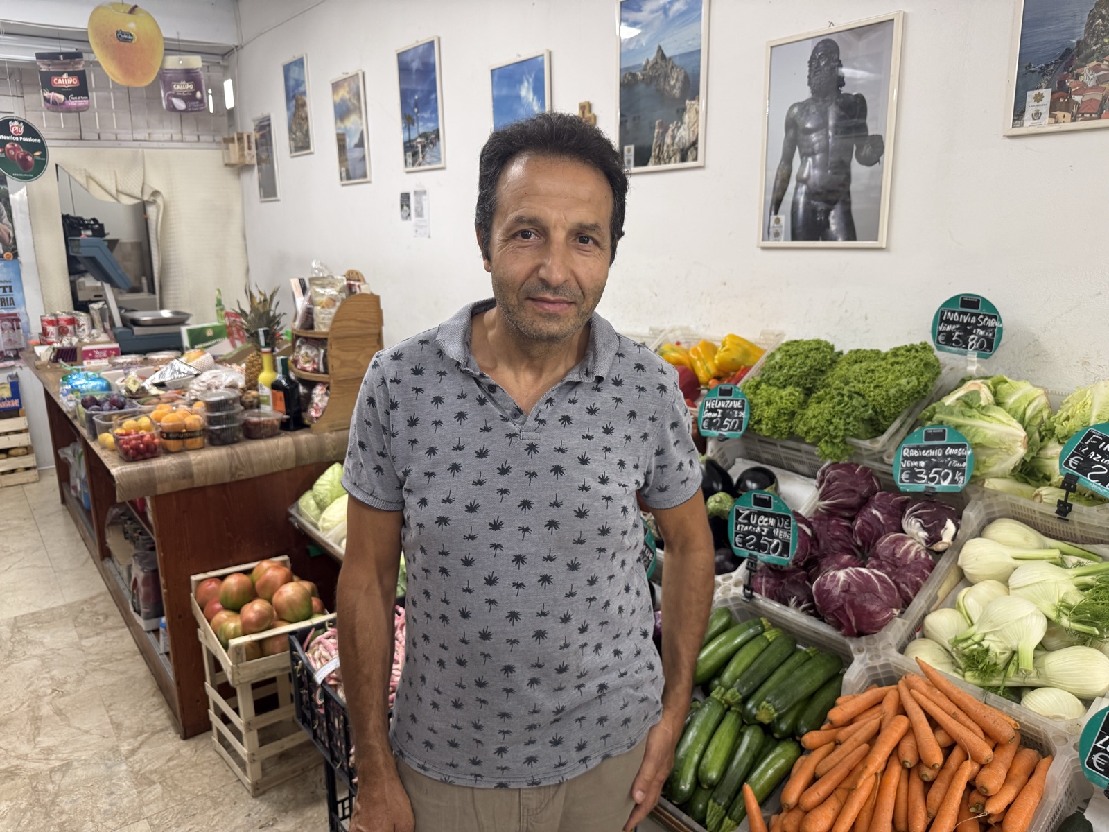 Antonino nella bottega di Via Fratelli Perini, tra il banco della verdura e le fotografie della Calabria, con i Bronzi di Riace alle pareti